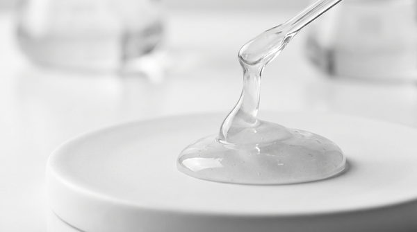 Flacone contagocce di siero senza etichetta accanto a una goccia di siero trasparente su una superficie in pietra chiara, still-life skincare luminoso e pulito.
