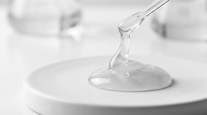 Flacone contagocce di siero senza etichetta accanto a una goccia di siero trasparente su una superficie in pietra chiara, still-life skincare luminoso e pulito.