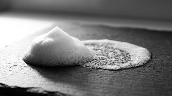Doppia detersione uomo: macro in bianco e nero di schiuma detergente che si scioglie su una lastra di ardesia bagnata, stile lab monocromo HBSKN